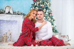 Loving Couple Husband And Wife In Plaid Pajamas Are Sitting Near The New Year Tree By The Fireplace And Hugging. Love And A Happy Royalty Free Stock Photo Loving Couple Husband And Wife In Plaid Pajamas Are Sitting Near The New Year Tree By The Fireplace And Hugging. Love And A Happy Royalty Free Stock Photo