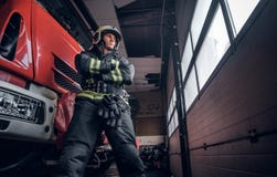 Low angle photo. Confident fireman wearing protective uniform standing next to a fire engine in a garage of a fire stock image Low angle photo. Confident fireman wearing protective uniform standing next to a fire engine in a garage of a fire stock image