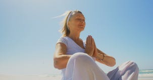 Low angle view of active senior Caucasian woman performing yoga on the beach 4k stock video Low angle view of active senior Caucasian woman performing yoga on the beach 4k stock video