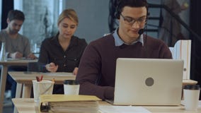 Lycklig gladlynt ung arbetare av appellmitten i huvud-telefoner som ler och arbetar på bärbara datorn arkivbild Lycklig gladlynt ung arbetare av appellmitten i huvud-telefoner som ler och arbetar på bärbara datorn arkivbild