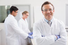 Lächelnder Wissenschaftler, der Kamera während Kollegen hinten arbeiten betrachtet stockfotografie Lächelnder Wissenschaftler, der Kamera während Kollegen hinten arbeiten betrachtet stockfotografie