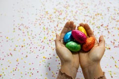 M?os da mulher que guardam ovos da p?scoa coloridos do chocolate com fundo branco e confetes borrados coloridos imagem de stock M?os da mulher que guardam ovos da p?scoa coloridos do chocolate com fundo branco e confetes borrados coloridos imagem de stock