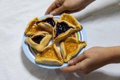 M?os da mulher que guardam uma placa colorida com as cookies do doce do mirtilo e do abric? de Hamantash Purim na toalha de mesa fotografia de stock M?os da mulher que guardam uma placa colorida com as cookies do doce do mirtilo e do abric? de Hamantash Purim na toalha de mesa fotografia de stock