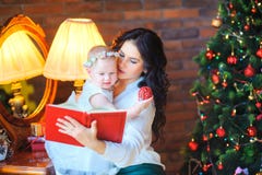 Madre che si siede vicino allo specchio che tiene una piccola figlia, sui precedenti di un albero di Natale festivo immagini stock Madre che si siede vicino allo specchio che tiene una piccola figlia, sui precedenti di un albero di Natale festivo immagini stock