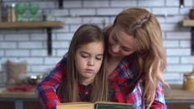 Madre joven hermosa que lee un libro para una pequeña hija linda que se sienta en la tabla de cocina El niño está trastornado y almacen de metraje de vídeo Madre joven hermosa que lee un libro para una pequeña hija linda que se sienta en la tabla de cocina El niño está trastornado y almacen de metraje de vídeo