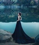 A majestic lady, a dark queen, stands on the background of a river and rocks, in a long black dress. The brunette girl