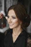 Makeup artist applying makeup on face of a beautiful and cheerful brunette model girl with charming smile in black Stock Photo Makeup artist applying makeup on face of a beautiful and cheerful brunette model girl with charming smile in black Stock Photo