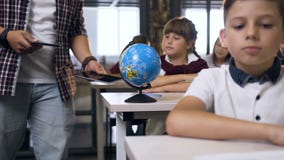 Male teacher of elementary school walking between desks puts computers tablet on desk pupils for learning