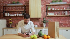 Man browsing on mobile phone at home kitchen. Handsome young man browsing on smartphone smiling happy. A man eats an stock footage Man browsing on mobile phone at home kitchen. Handsome young man browsing on smartphone smiling happy. A man eats an stock footage