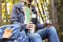 Man with cup of hot drink - tea or coffee and thermos sits in autumn forest with backpack, maple leaf stock photo Man with cup of hot drink - tea or coffee and thermos sits in autumn forest with backpack, maple leaf stock photo