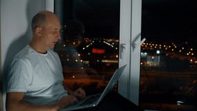Man genom att använda anteckningsboksammanträde på fönsterbräda med panoramautsikt på nattcityscape lager videofilmer Man genom att använda anteckningsboksammanträde på fönsterbräda med panoramautsikt på nattcityscape lager videofilmer