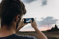 man hipster traveler taking photo holding smart phone, of beautiful sunset scenery in summer field. instagram photography. royalty free stock images man hipster traveler taking photo holding smart phone, of beautiful sunset scenery in summer field. instagram photography. royalty free stock images