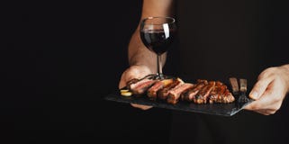 Man holding juicy grilled beef steak with spices and red wine glass on a stone cutting board on a black background. With copy spac
