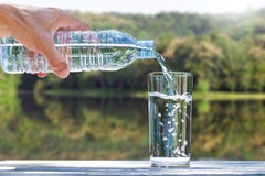 Man& x27;s hand holding drinking bottle water and pouring water into glass on wooden table on blurred green nature background stock photo Man& x27;s hand holding drinking bottle water and pouring water into glass on wooden table on blurred green nature background stock photo