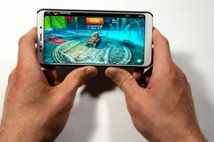 Mani che tengono uno smartphone su cui il gioco, mondo gameplay di Gameplay di attacco dei carri armati immagine stock libera da diritti Mani che tengono uno smartphone su cui il gioco, mondo gameplay di Gameplay di attacco dei carri armati immagine stock libera da diritti