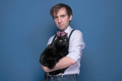 Mann im Hemd und in rosa Fliege, die nette lustige schwarze Katze halten und Kamera auf blauem Hintergrund betrachten lizenzfreie stockbilder Mann im Hemd und in rosa Fliege, die nette lustige schwarze Katze halten und Kamera auf blauem Hintergrund betrachten lizenzfreie stockbilder