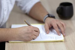 The mans hands on white outfit writes with roller pen on the paper on wooden table some latin, or medicine terms. The cup of tea stock photography The mans hands on white outfit writes with roller pen on the paper on wooden table some latin, or medicine terms. The cup of tea stock photography