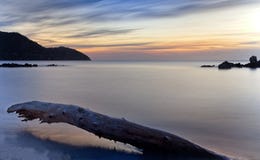 Mare liscio ad alba, con il cielo, le colline, le rocce ed il tronco di albero commoventi drammatici sulla spiaggia, nou del port immagini stock Mare liscio ad alba, con il cielo, le colline, le rocce ed il tronco di albero commoventi drammatici sulla spiaggia, nou del port immagini stock