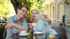 Blonde taking selfie with friend in outdoor cafe holding coffee using smartphone