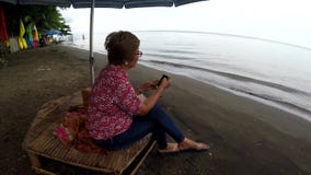 Mature woman browses internet with the use of mobile smartphone while sitting cross legged under parasol sunshade stock footage Mature woman browses internet with the use of mobile smartphone while sitting cross legged under parasol sunshade stock footage