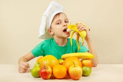 Mała śmieszna chłopiec je świeżego banana przy stołem z owoc zdjęcia royalty free Mała śmieszna chłopiec je świeżego banana przy stołem z owoc zdjęcia royalty free