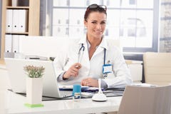 Medico femminile felice che si siede alla scrivania medica fotografia stock Medico femminile felice che si siede alla scrivania medica fotografia stock