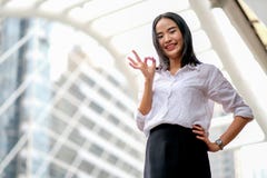 Menina bonita asiática do negócio com ato branco da camisa como o sinal seguro e da mostra da APROVAÇÃO na cidade grande no tempo imagem de stock Menina bonita asiática do negócio com ato branco da camisa como o sinal seguro e da mostra da APROVAÇÃO na cidade grande no tempo imagem de stock