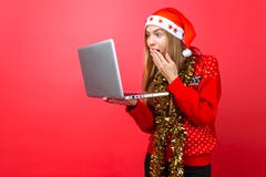 A menina chocada em uma camiseta e em um chapéu vermelhos de Santa Claus, com um portátil à disposição, olha na surpresa na tela fotos de stock A menina chocada em uma camiseta e em um chapéu vermelhos de Santa Claus, com um portátil à disposição, olha na surpresa na tela fotos de stock