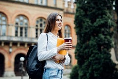 Menina de sorriso feliz do estudante com a trouxa no fundo de universidade A mulher com manuais de instruções e leva embora o caf fotos de stock Menina de sorriso feliz do estudante com a trouxa no fundo de universidade A mulher com manuais de instruções e leva embora o caf fotos de stock