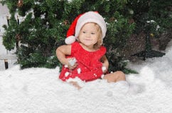 A menina de sorriso no traje de Santa senta-se na neve imagens de stock royalty free A menina de sorriso no traje de Santa senta-se na neve imagens de stock royalty free