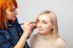A menina do maquilhador com cabelo vermelho e uma escova em sua mão impõe a composição nos olhos do modelo - máscaras da cor dour fotos de stock royalty free A menina do maquilhador com cabelo vermelho e uma escova em sua mão impõe a composição nos olhos do modelo - máscaras da cor dour fotos de stock royalty free