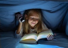 Menina feliz bonita que lê um livro na escuridão que esconde sob a edredão na cama com luz instantânea fotos de stock Menina feliz bonita que lê um livro na escuridão que esconde sob a edredão na cama com luz instantânea fotos de stock