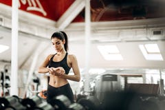 A menina magro bonita vestida no sportswear preto está estando no gym e está olhando seu relógio imagens de stock A menina magro bonita vestida no sportswear preto está estando no gym e está olhando seu relógio imagens de stock