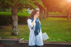 Menina moreno bonita em um lenço na rua que fala no telefone que guarda um portátil imagem de stock Menina moreno bonita em um lenço na rua que fala no telefone que guarda um portátil imagem de stock