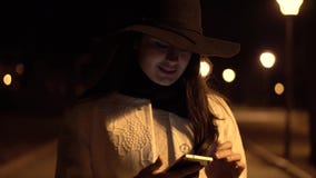 A menina moreno nova em um chapéu anda no parque da noite e começa verificar seu telefone video estoque A menina moreno nova em um chapéu anda no parque da noite e começa verificar seu telefone video estoque
