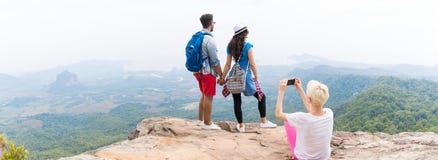 Menina que toma a foto dos pares com as trouxas que levantam sobre a paisagem da montanha no panorama esperto do telefone da pilh imagem de stock royalty free Menina que toma a foto dos pares com as trouxas que levantam sobre a paisagem da montanha no panorama esperto do telefone da pilh imagem de stock royalty free