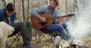 Mens het spelen gitaar en vriend het strijken hond samen rond brandplaats Echte vriendenmensen openlucht het kamperen tentvakanti stock video Mens het spelen gitaar en vriend het strijken hond samen rond brandplaats Echte vriendenmensen openlucht het kamperen tentvakanti stock video