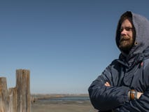 Mens hipster met een baard en een snor in de kap tegen de blauwe hemel en het estuarium royalty-vrije stock foto's Mens hipster met een baard en een snor in de kap tegen de blauwe hemel en het estuarium royalty-vrije stock foto's