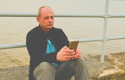 Midle a vieilli l'homme s'asseyant sur le rivage du lac Homme mûr de solitude à l'aide du téléphone portable sur la banque Concep Image libre de droits Midle a vieilli l'homme s'asseyant sur le rivage du lac Homme mûr de solitude à l'aide du téléphone portable sur la banque Concep Image libre de droits