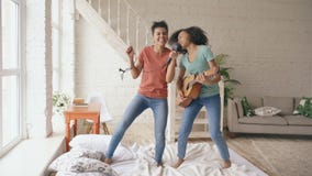 Mixed race young funny girls dance singing with hairdryer and playing acoustic guitar on a bed. Sisters having fun