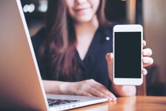 Mockup image of a smiley Asian beautiful woman holding and showing white mobile phone with blank black screen while using laptop