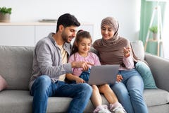 Modern muslim parents teaching their daughter using laptop computer, sitting together on sofa at home