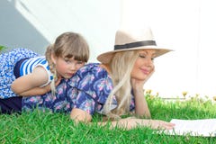 Mom blonde in a hat and her daughter are lying on the grass and reading a book, happy family. stock photo Mom blonde in a hat and her daughter are lying on the grass and reading a book, happy family. stock photo