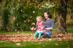 Mother And Girl Having Fun Under Tree With Autumn Leaves In The Park, Blond Cute Curly Girl, Happy And Young Family Stock Photo