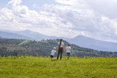 Mother and two little sons stand holding hands on a green field against a background of forest, mountains and sky with clouds stock photo Mother and two little sons stand holding hands on a green field against a background of forest, mountains and sky with clouds stock photo