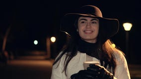 A moça moreno em um chapéu e em um revestimento branco bebe o café e sorri em um parque da noite video estoque A moça moreno em um chapéu e em um revestimento branco bebe o café e sorri em um parque da noite video estoque