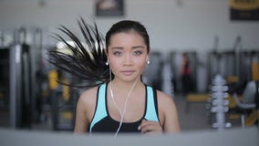 Muchacha asiática atractiva que corre en la rueda de ardilla en el gimnasio con los auriculares Retrato de la cara llena metrajes Muchacha asiática atractiva que corre en la rueda de ardilla en el gimnasio con los auriculares Retrato de la cara llena metrajes