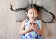Muchacha asiática del niño que mira el smartphone que miente en piso de madera Visión superior foto de archivo libre de regalías Muchacha asiática del niño que mira el smartphone que miente en piso de madera Visión superior foto de archivo libre de regalías