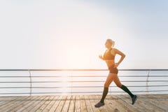 Muchacha atlética hermosa joven con el pelo rubio largo en la ropa negra que corre en la salida del sol sobre el mar Imágenes de archivo libres de regalías Muchacha atlética hermosa joven con el pelo rubio largo en la ropa negra que corre en la salida del sol sobre el mar Imágenes de archivo libres de regalías