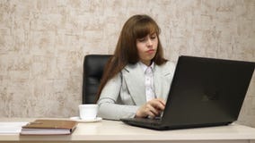 Muchacha hermosa que se sienta en silla en oficina y que mecanografía en el ordenador portátil y el café de consumición de la taz fotos de archivo Muchacha hermosa que se sienta en silla en oficina y que mecanografía en el ordenador portátil y el café de consumición de la taz fotos de archivo